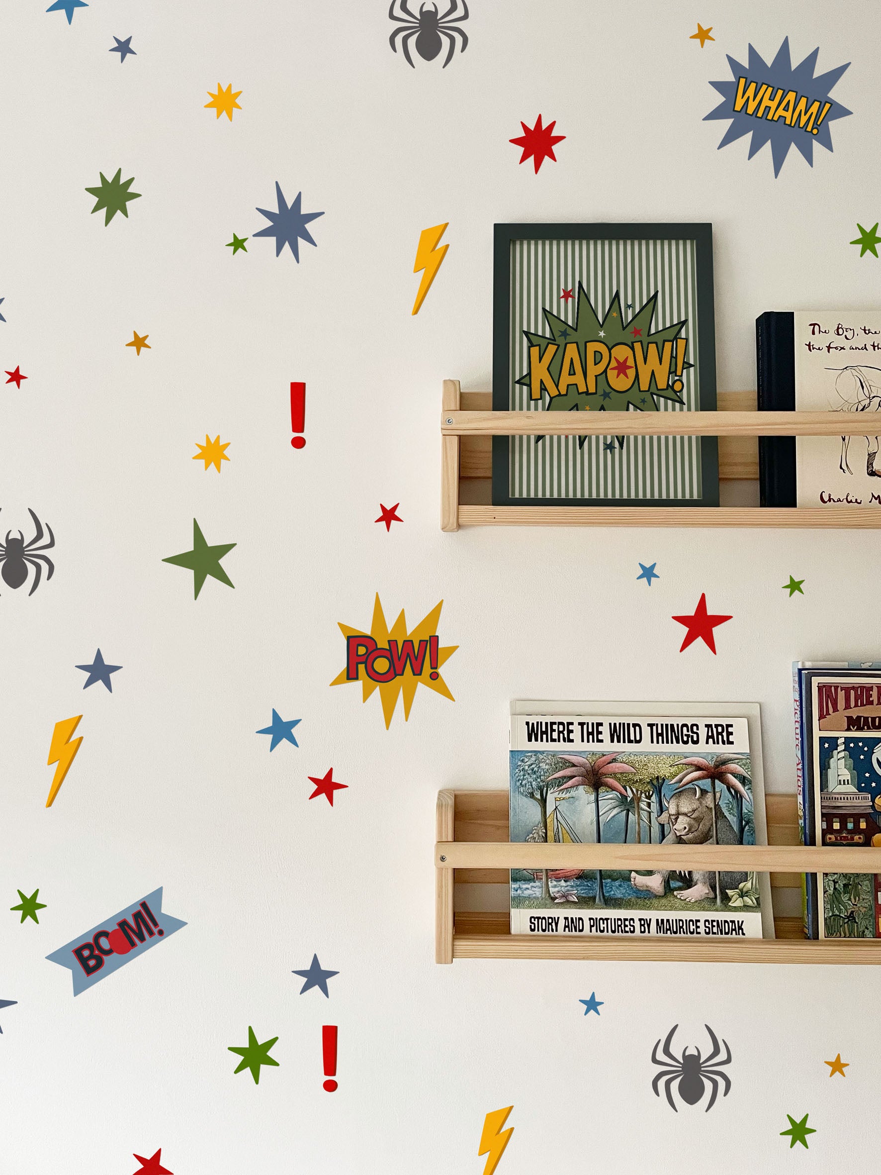 A white wall decorated with colorful comic book themed wall decals, including stars, lightning bolts, exclamation marks, spider silhouettes, and burst shapes with words like 'WHAM!', 'BOOM!', and 'KAPOW!'. Two light wood floating shelves are mounted on the wall, holding a framed print and children's books.