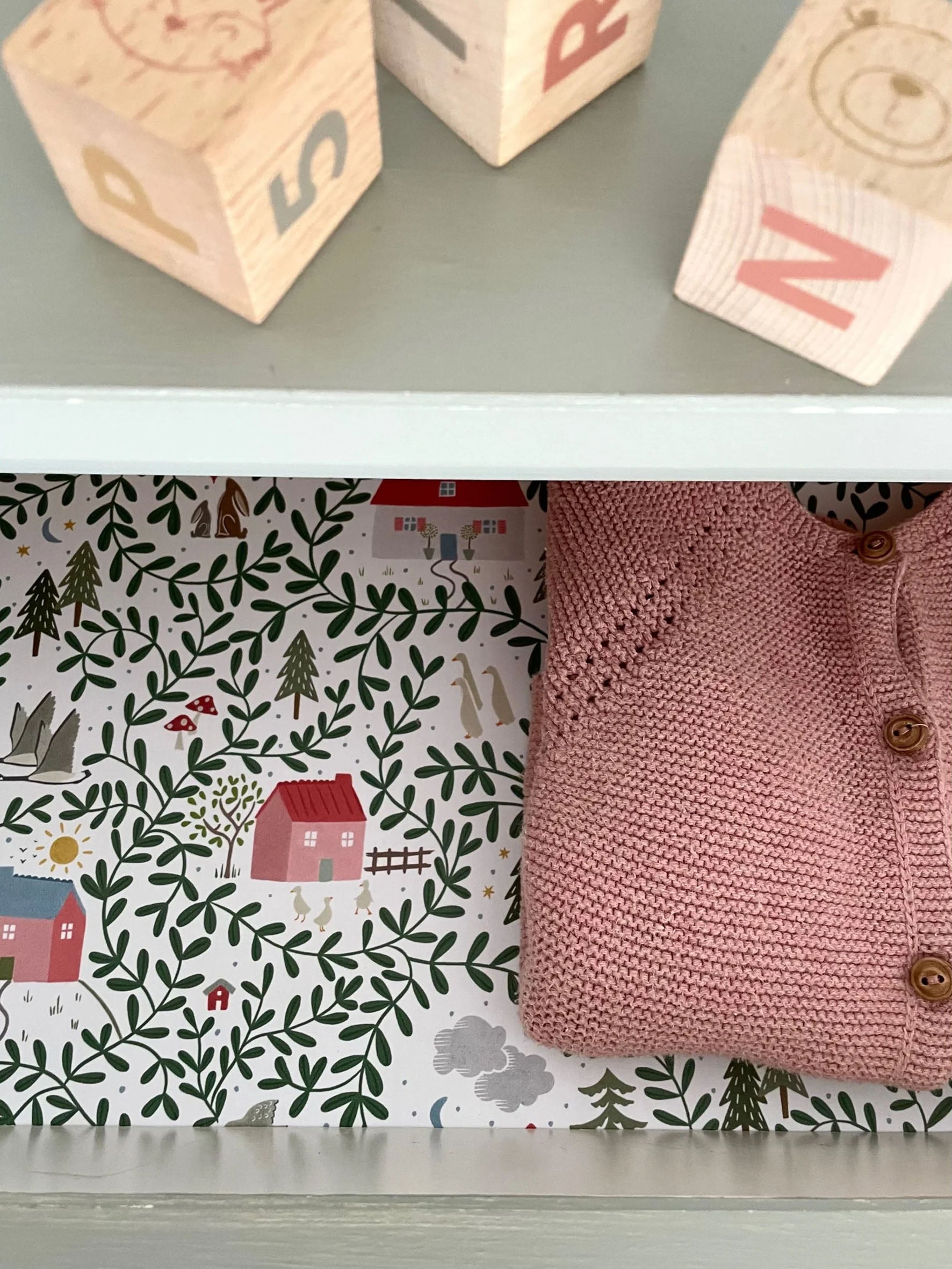 Childrens chest of drawers lined with english country cottages wallpaper, with building blocks and pink baby girls cardigan